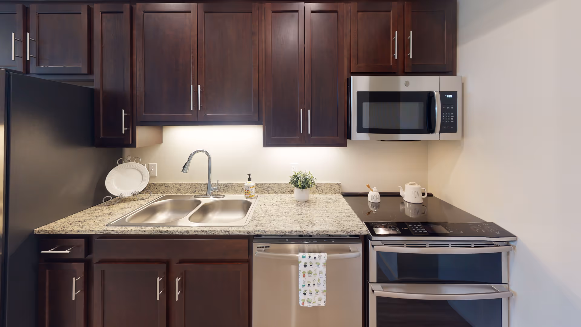 A modern kitchen with dark wooden cabinets, a granite countertop, a double stainless steel sink with a chrome faucet, a stainless steel dishwasher with a decorative towel hanging on the handle, a stainless steel electric stove with two ovens, and a stainless steel microwave mounted above the stove. There is a white decorative plate on a stand, a small potted plant, and two ceramic containers labeled 'Honey' and 'Tea' on the countertop.