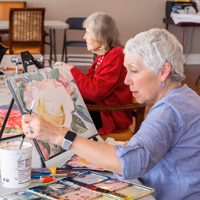 Two elderly women seated indoors, one in the foreground painting a large flower on a canvas with watercolors, and the other in the background wearing a red sweater. Various painting supplies are on the table in front of them.