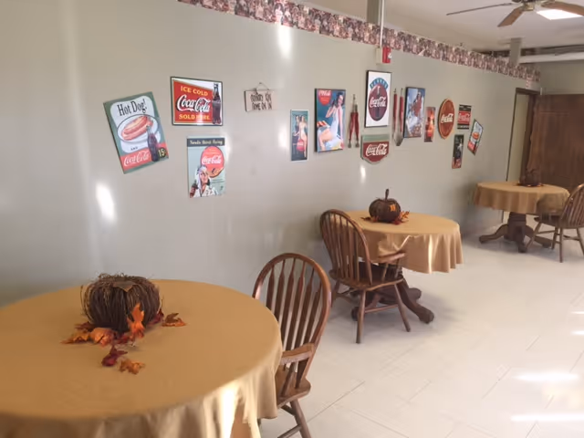 Small dining area with round tables covered in beige tablecloths, wooden chairs, and vintage Coca-Cola wall signs.