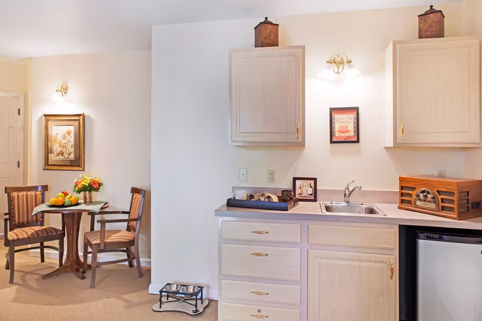 A cozy interior space featuring a small dining area with a round wooden table and two striped cushioned chairs. The table has a vase with flowers and a bowl of fruit. Adjacent to the dining area is a kitchenette with light wood cabinets, a countertop with a sink, a vintage-style radio, and a mini refrigerator. The walls are decorated with framed artwork and soft lighting fixtures. A pet feeding station with two bowls is on the floor near the kitchenette.