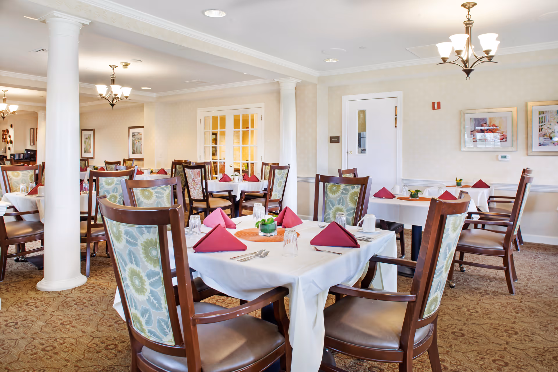 A bright and elegant dining room with multiple tables covered in white tablecloths, each set with red folded napkins, glasses, and silverware. The chairs have wooden frames with floral-patterned upholstery. The room features beige walls, carpeted floors, white columns, and framed artwork on the walls. Ceiling lights with multiple bulbs provide warm lighting.