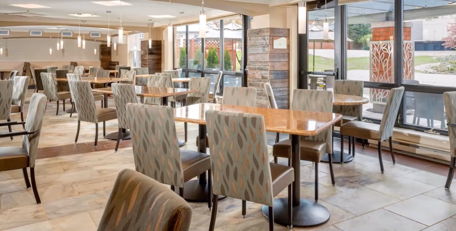A spacious dining room with multiple tables and patterned upholstered chairs arranged neatly. Large windows on one side allow natural light to fill the room, and pendant lights hang from the ceiling. The floor is tiled, and there are wooden accent walls near the windows.