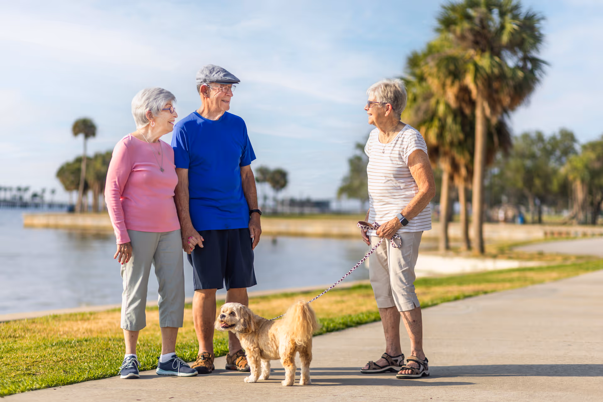 Three elderly people standing on a paved path near a body of water with palm trees in the background. One woman is holding a small dog on a leash, and the other two people are standing close together, smiling and talking.
