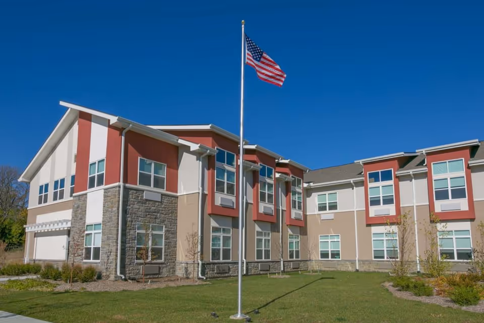 Two-story modern building with red and beige siding and an American flag on a flagpole on a grassy lawn under a clear blue sky.