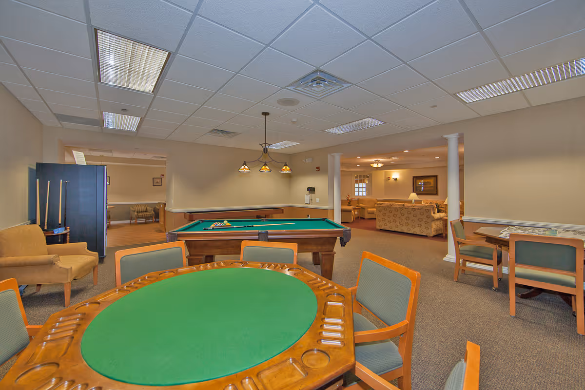 A spacious recreational room in a senior living facility featuring a green felt poker table with wooden chairs in the foreground, a pool table with cues and balls in the middle, and a cozy seating area with sofas and armchairs in the background. The room has beige walls, carpeted floors, and a drop ceiling with fluorescent lighting.