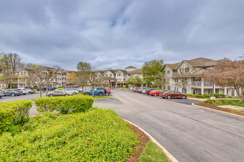 Exterior view of a senior living facility with a large parking lot filled with cars, surrounded by green bushes and trees under a cloudy sky.