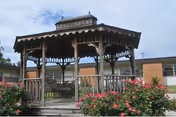 A wooden gazebo with a shingled roof situated in a garden area with blooming pink flowers and green bushes, surrounded by a single-story building under a partly cloudy sky.