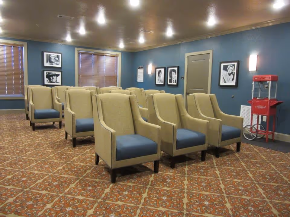 A small theater or screening room with three rows of beige armchairs with blue cushions arranged facing forward. The room has blue walls adorned with black and white framed portraits and two windows with closed blinds. A red popcorn machine is positioned in the corner near a closed door. The floor is covered with a patterned carpet.