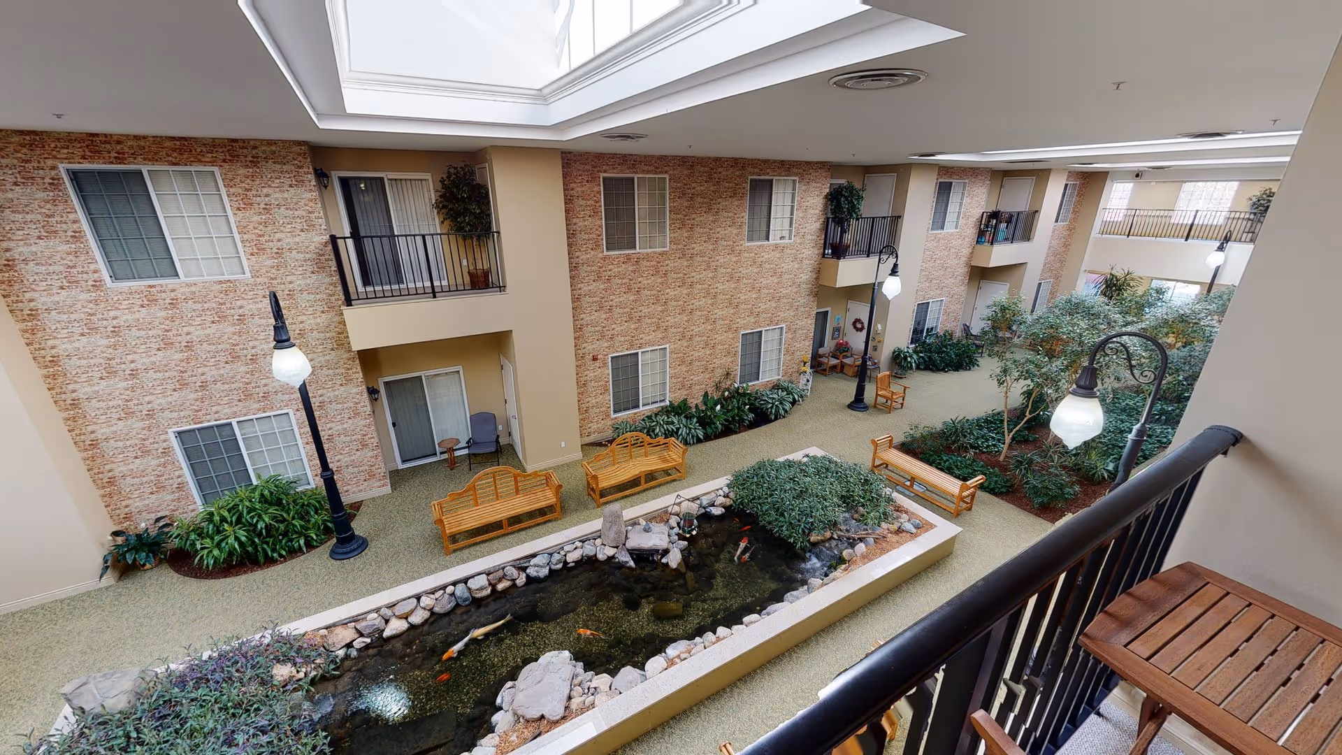 Indoor courtyard area of a senior living facility with a koi pond surrounded by rocks and plants. There are wooden benches and chairs placed around the pond and along the walkway. The walls have a brick pattern with several windows and doors leading to rooms. The ceiling has skylights allowing natural light to illuminate the space. Black metal railings and vintage-style lamp posts add to the decor.