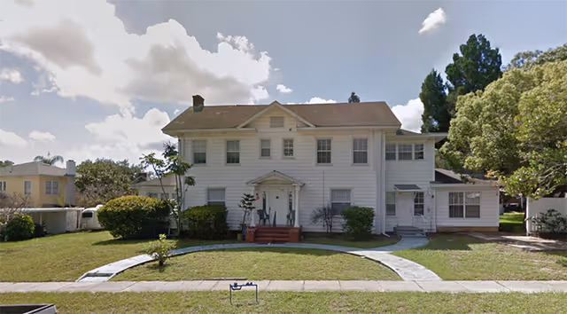 Front exterior view of a two-story white house with a brown roof, surrounded by green grass, bushes, and trees under a partly cloudy sky.