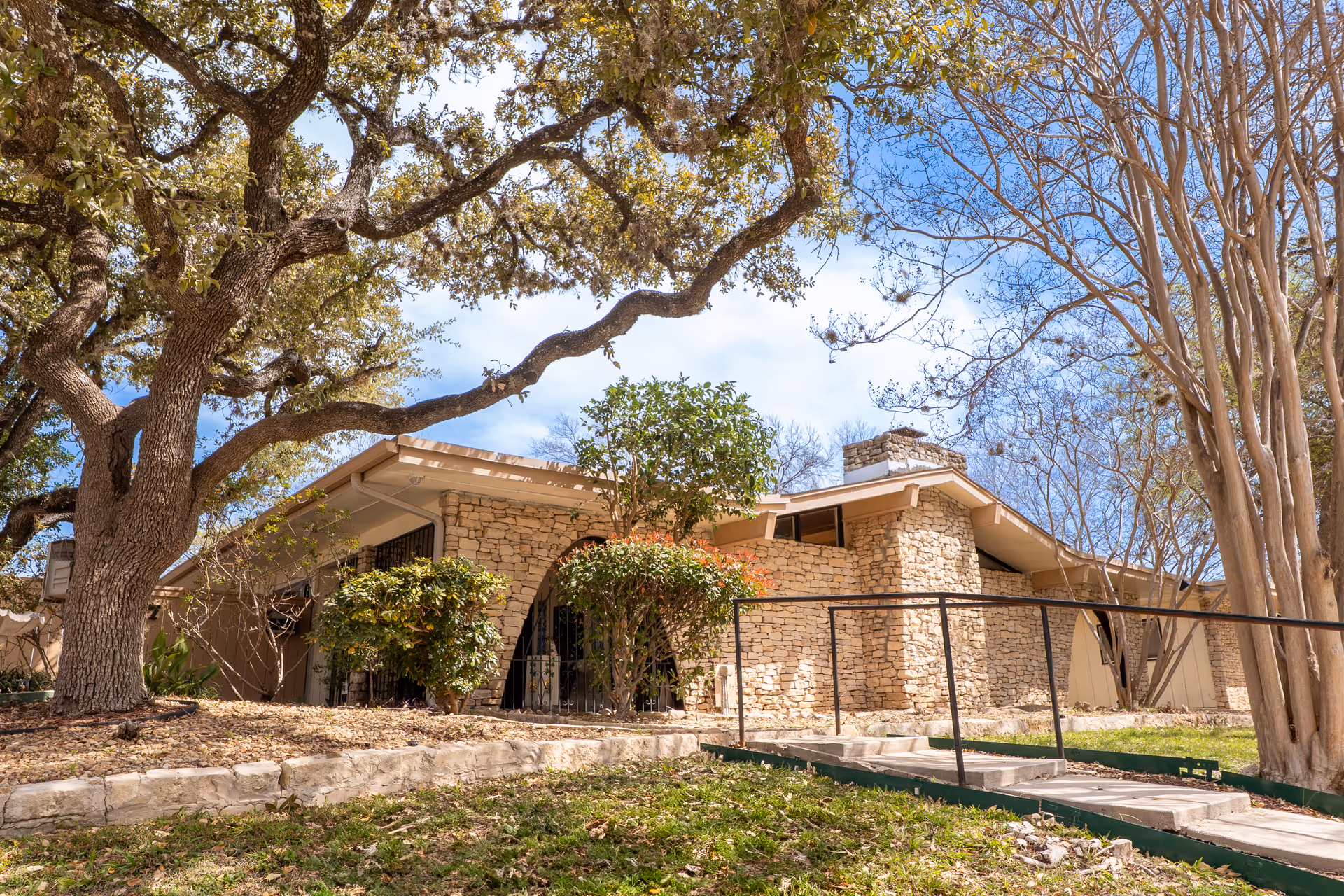 Stone building with a sloped roof surrounded by trees and bushes under a blue sky with some clouds. A ramp with a black handrail leads up to the entrance.