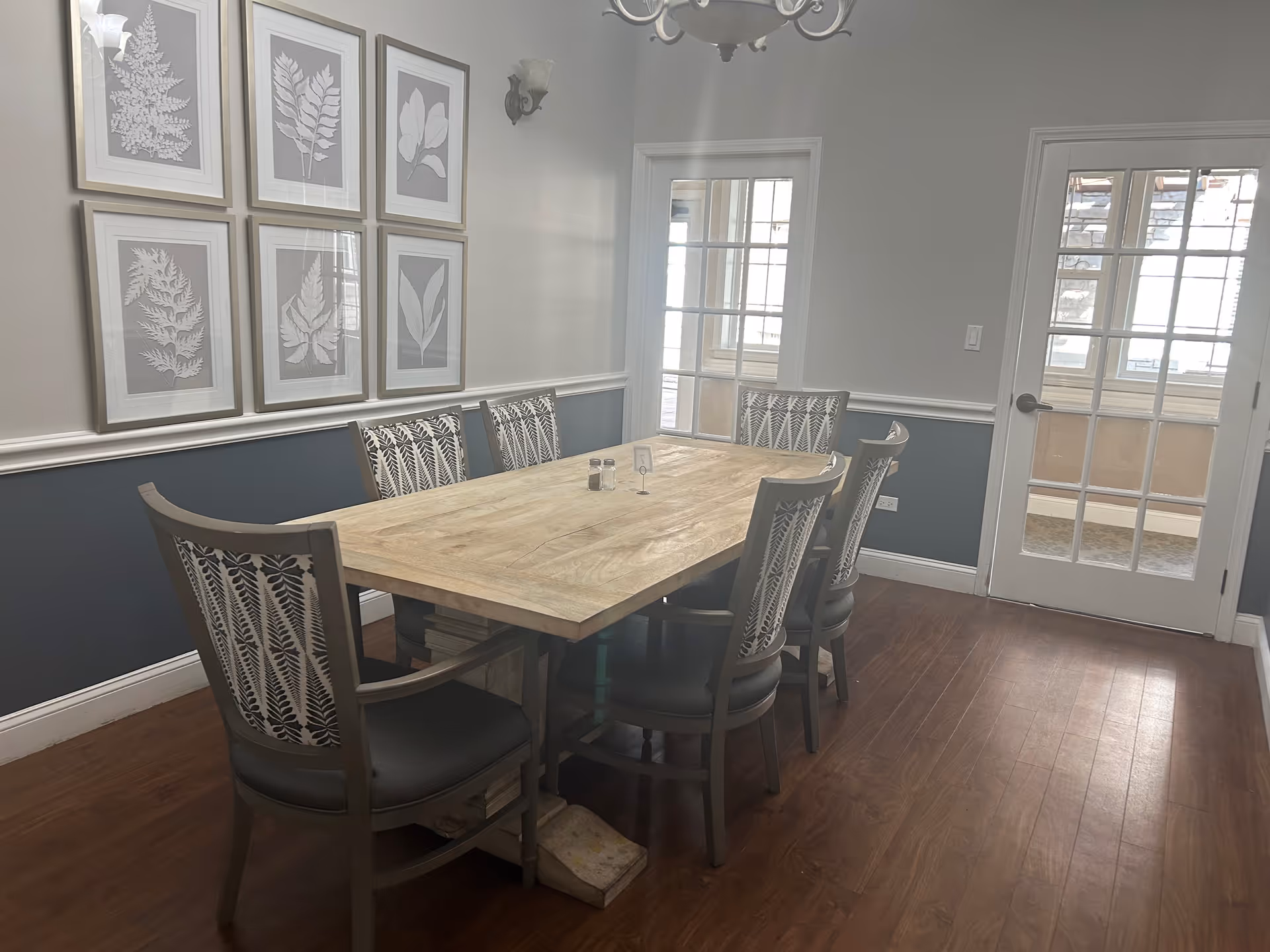 A dining room with a wooden rectangular table surrounded by six chairs with patterned backs. The walls are painted gray with white trim and decorated with six framed botanical prints. There are two wall sconces and a chandelier above the table. Two glass-paneled doors are visible, allowing natural light into the room.