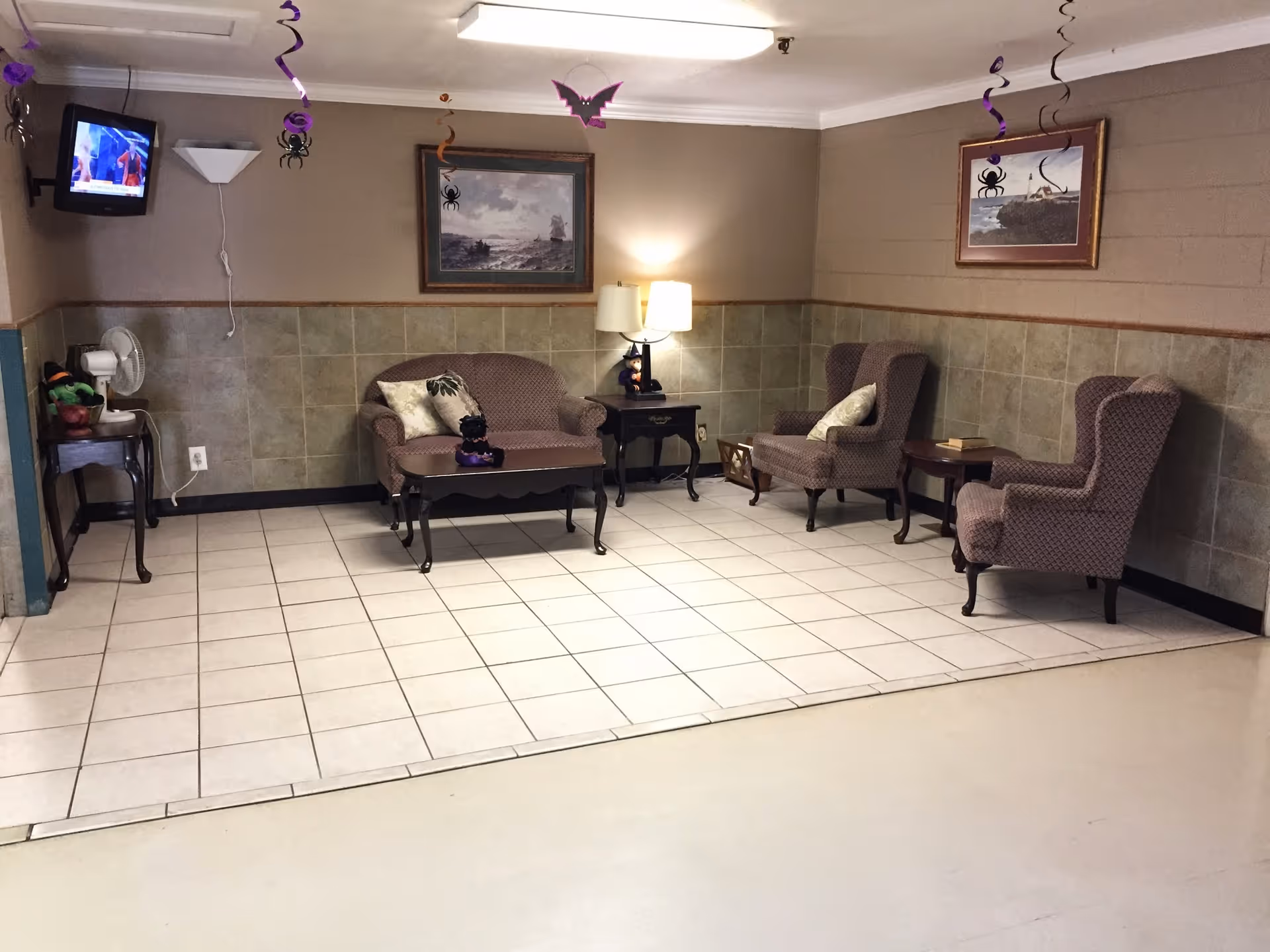 A small sitting area in a nursing home decorated for Halloween with hanging purple and orange streamers, paper bats, and spiders. The room has a tiled floor section with a loveseat, two armchairs, a coffee table, and two side tables with lamps. There are framed paintings on the walls and a small TV mounted in the corner.