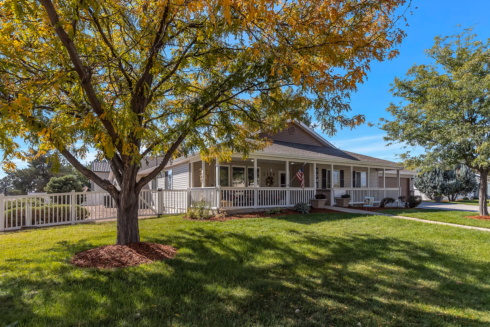 Single-story memory care building with a wide front porch, white railing, American flag, and trees on a sunny day.