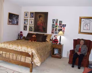 A cozy bedroom with a large bed, nightstand lamp, framed portraits, and a seated woman.
