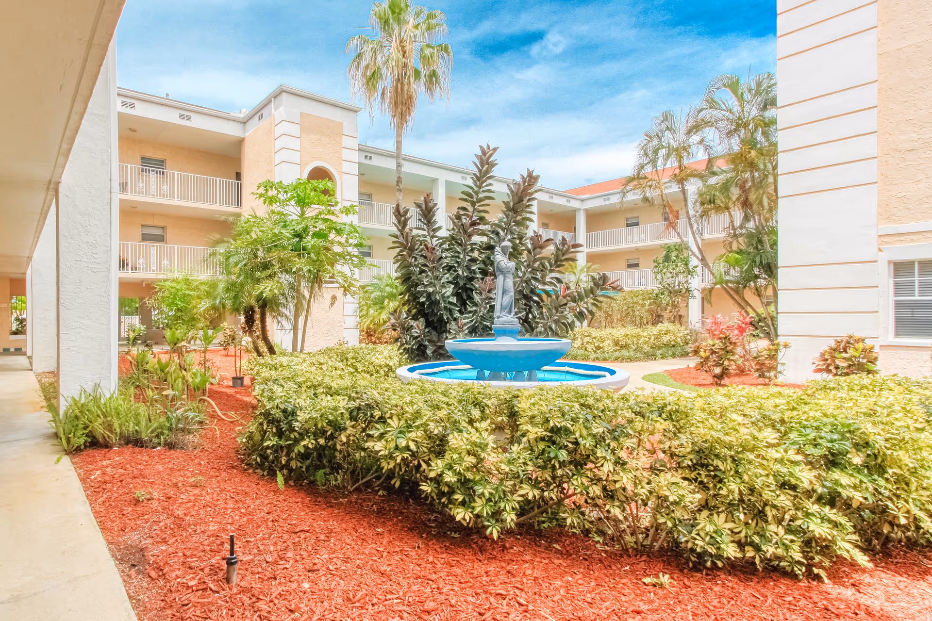 Outdoor courtyard area of a senior living facility with a central blue fountain surrounded by green shrubs and red mulch. The courtyard is enclosed by a three-story beige building with balconies and palm trees under a blue sky.