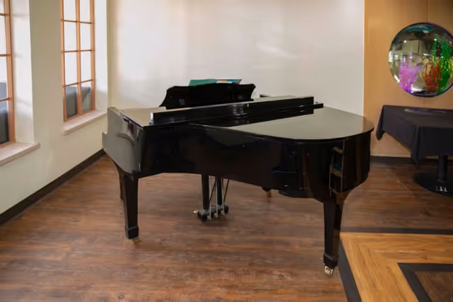 A black grand piano placed on a wooden floor in a room with light-colored walls and windows on the left side. There is a round mirror on the right wall reflecting colorful decorations and a table covered with a black cloth nearby.