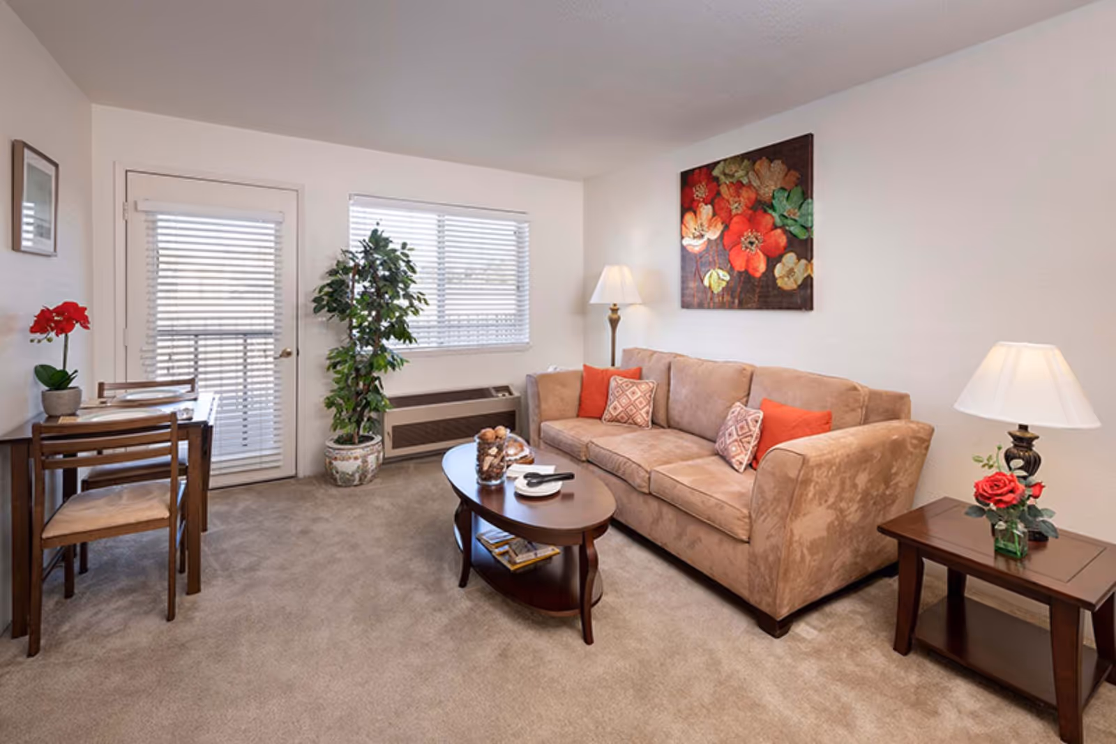 A cozy living room with a beige couch adorned with red and patterned pillows, a wooden coffee table with decorative items, a side table with a lamp and a red rose in a vase, a small dining table with two chairs and place settings, a potted plant near a window with blinds, and a door with blinds leading outside. A colorful floral painting hangs on the wall above the couch.