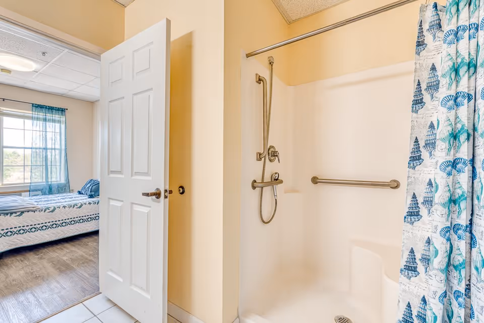 View of a bathroom shower with grab bars and a seashell-patterned shower curtain, adjacent to an open door leading to a bedroom with a bed and a window with blue curtains.
