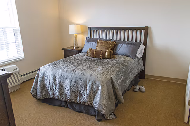 A neatly made bed with a gray patterned comforter and multiple pillows in a simple bedroom. There is a wooden headboard, a nightstand with a lamp, a window with blinds, and a pair of slippers on the floor next to the bed.