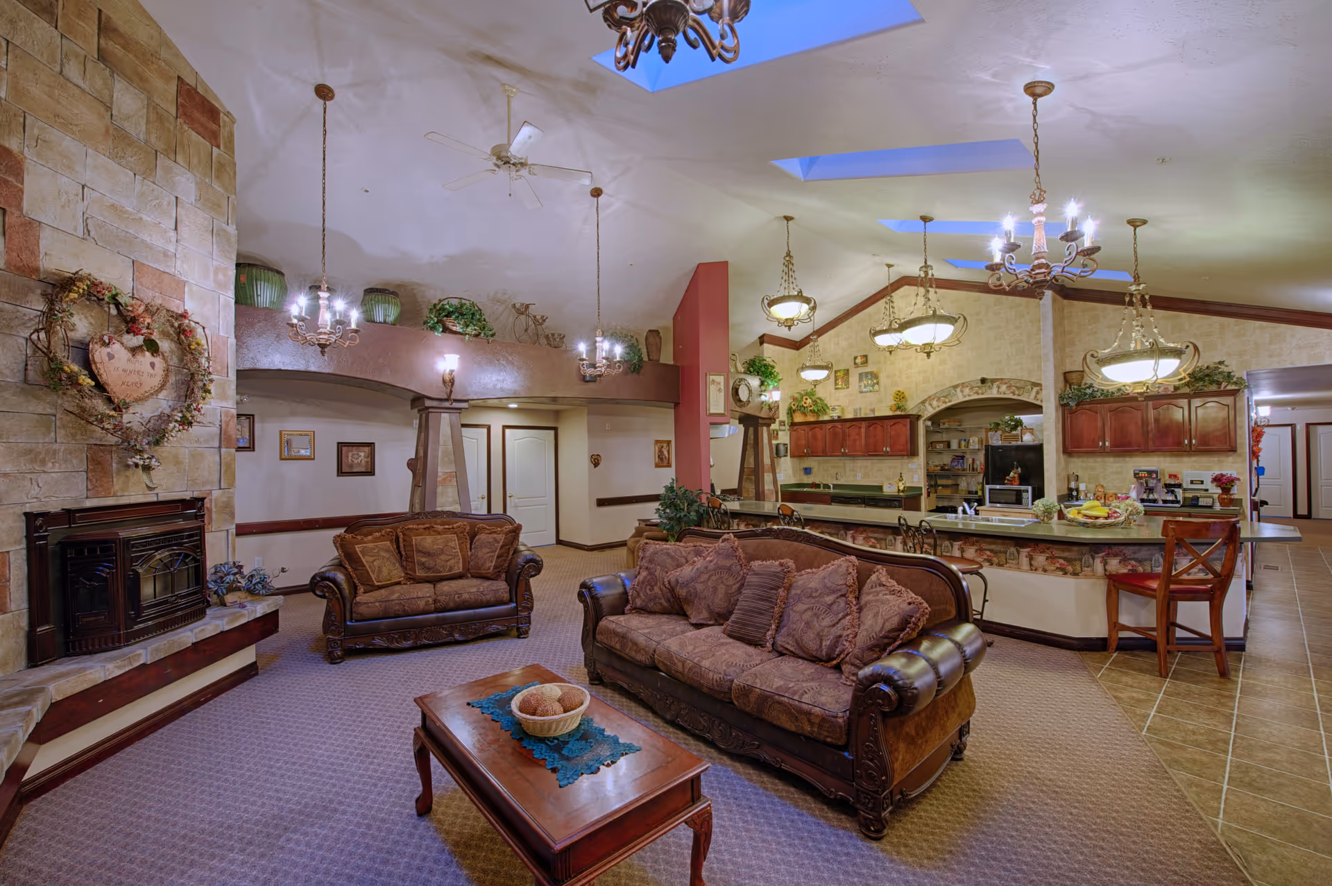 A spacious living room area with two brown patterned sofas, a wooden coffee table with a decorative bowl, and a stone fireplace adorned with a heart-shaped wreath. The room features multiple hanging light fixtures, ceiling fans, skylights, and an open view into a kitchen area with wooden cabinets and a breakfast bar with stools.