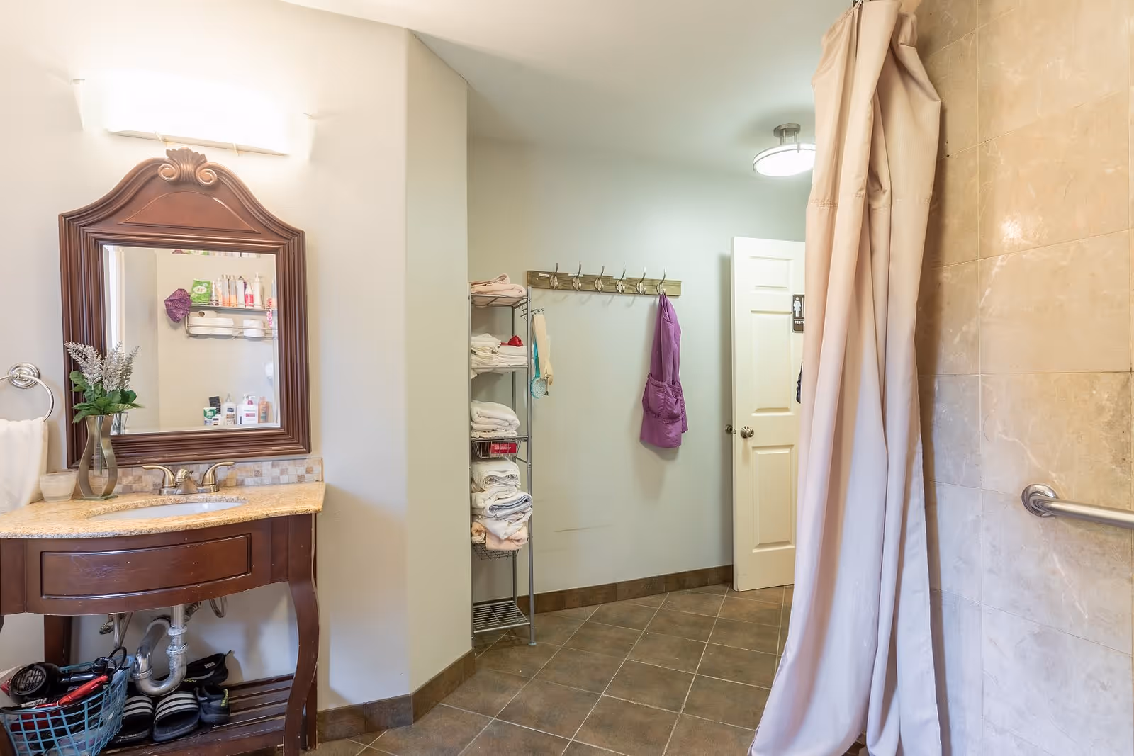 A bathroom with a wooden vanity topped with a granite countertop and a decorative mirror above it. There is a vase with flowers on the vanity. To the right, there is a shower area with a beige curtain and tiled walls. On the back wall, there is a metal rack holding folded towels and a wooden coat rack with a purple robe hanging on it. The floor is tiled in a brown pattern.