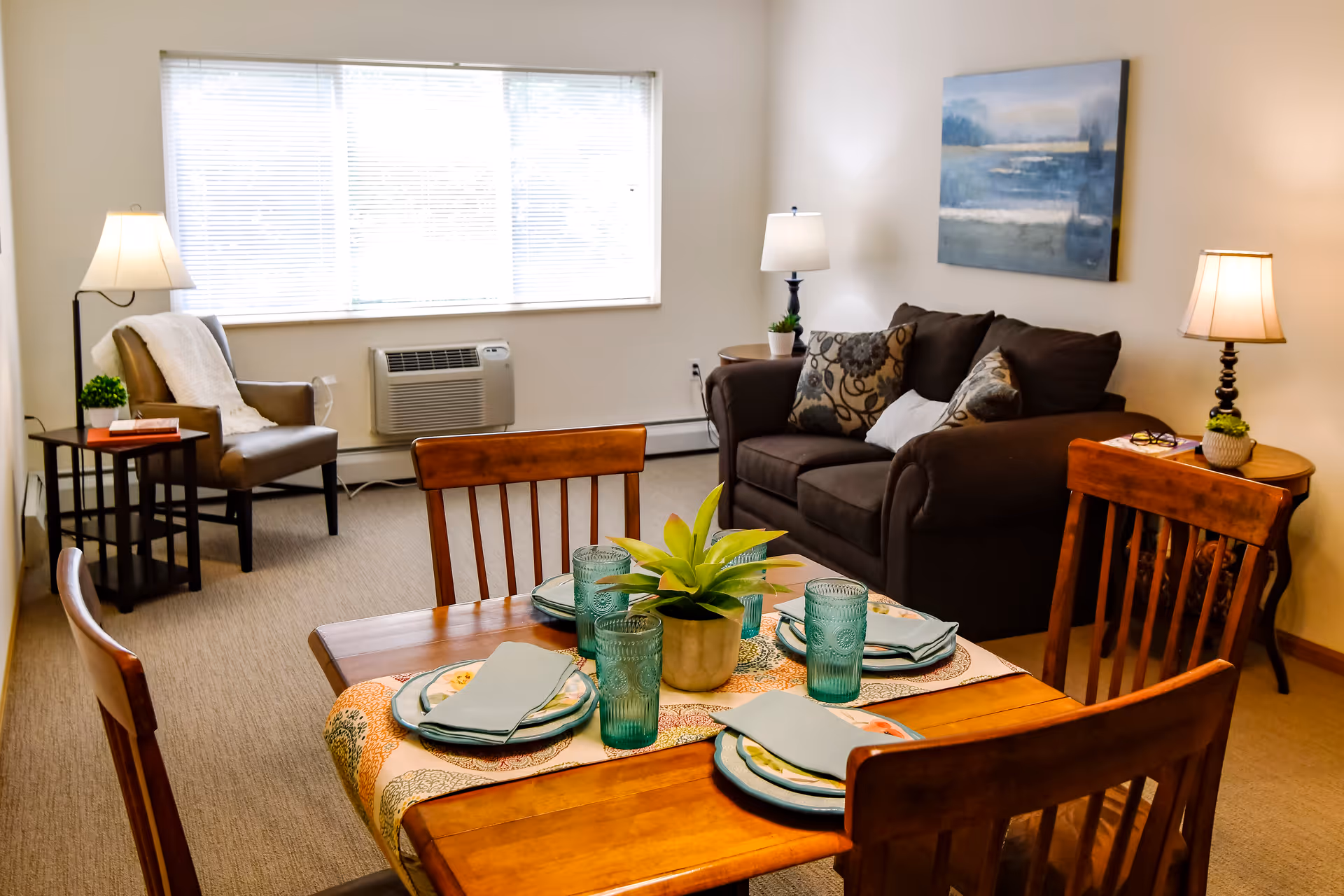 A cozy living and dining area with a wooden dining table set for four with plates, napkins, and blue glasses. In the background, there is a dark brown sofa with patterned cushions, a side table with a lamp and a small plant, and an armchair with a white throw blanket next to another lamp. A large window with blinds lets in natural light, and a blue-toned painting hangs on the wall above the sofa.