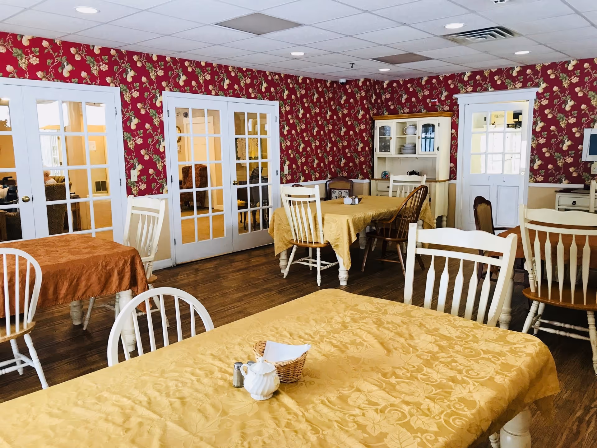 A dining room with several tables covered in yellow and orange tablecloths, surrounded by white and wooden chairs. The walls are decorated with red floral wallpaper, and there are white French doors and a white cabinet with dishes. The floor is wooden.