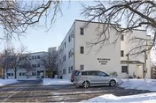 Exterior view of Wildwood Manor, a three-story senior living facility building with a parking lot in front. Snow covers the ground and leafless tree branches are visible in the foreground.