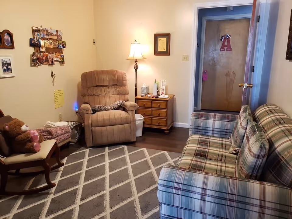A cozy living room with a plaid sofa on the right, a beige recliner chair in the center, and a wooden rocking chair with a teddy bear on the left. There is a patterned rug on the floor, a wooden side table with a lamp and various items, and a door in the background with a decorative letter 'A' hanging on it. The walls have framed pictures and a collage of photos.