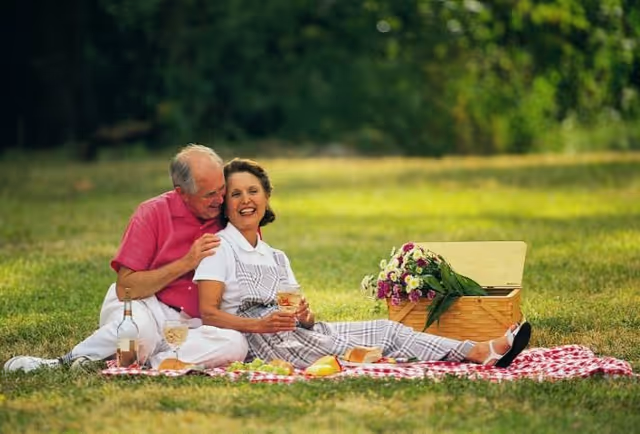 An elderly couple enjoying a picnic on a red and white checkered blanket in a grassy outdoor area. The man in a pink shirt sits behind the woman, who is wearing a white shirt and plaid overalls, holding a glass. There is a picnic basket with flowers and food items around them.