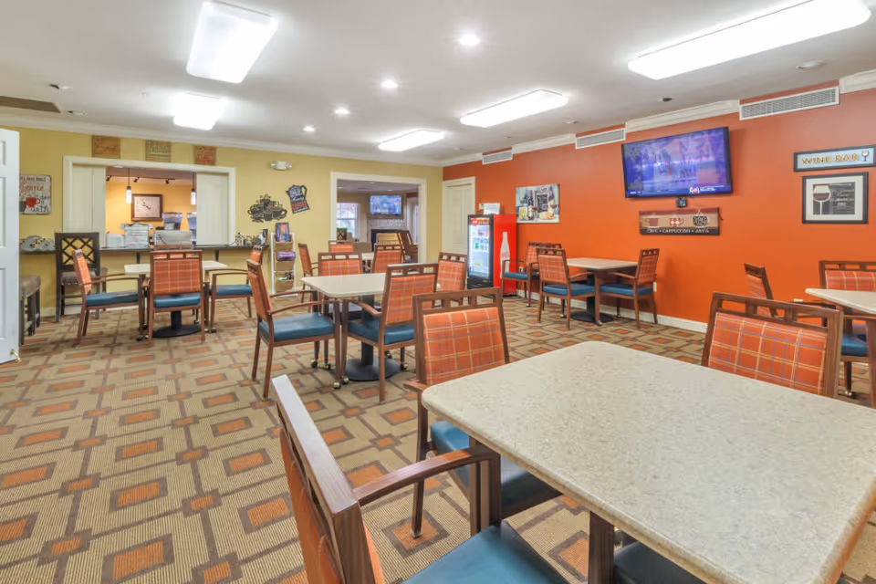 A bright communal dining room with multiple tables and chairs, patterned carpet, and an orange accent wall with a mounted TV and beverage fridge.