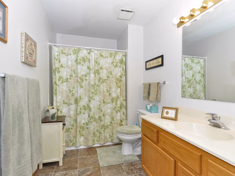 A clean bathroom with a floral green and white shower curtain, a toilet with a green cushioned seat cover, a wooden vanity with a white countertop and sink, a large mirror above the sink, green towels hanging on the wall, a small cabinet with a decorative plant, and framed wall art.