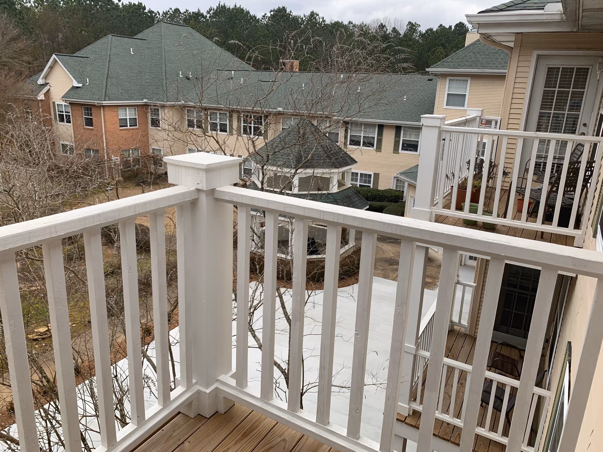 View from a balcony with white wooden railings overlooking a courtyard area and adjacent buildings with green roofs and beige walls. There are leafless trees and a gazebo-like structure in the courtyard.