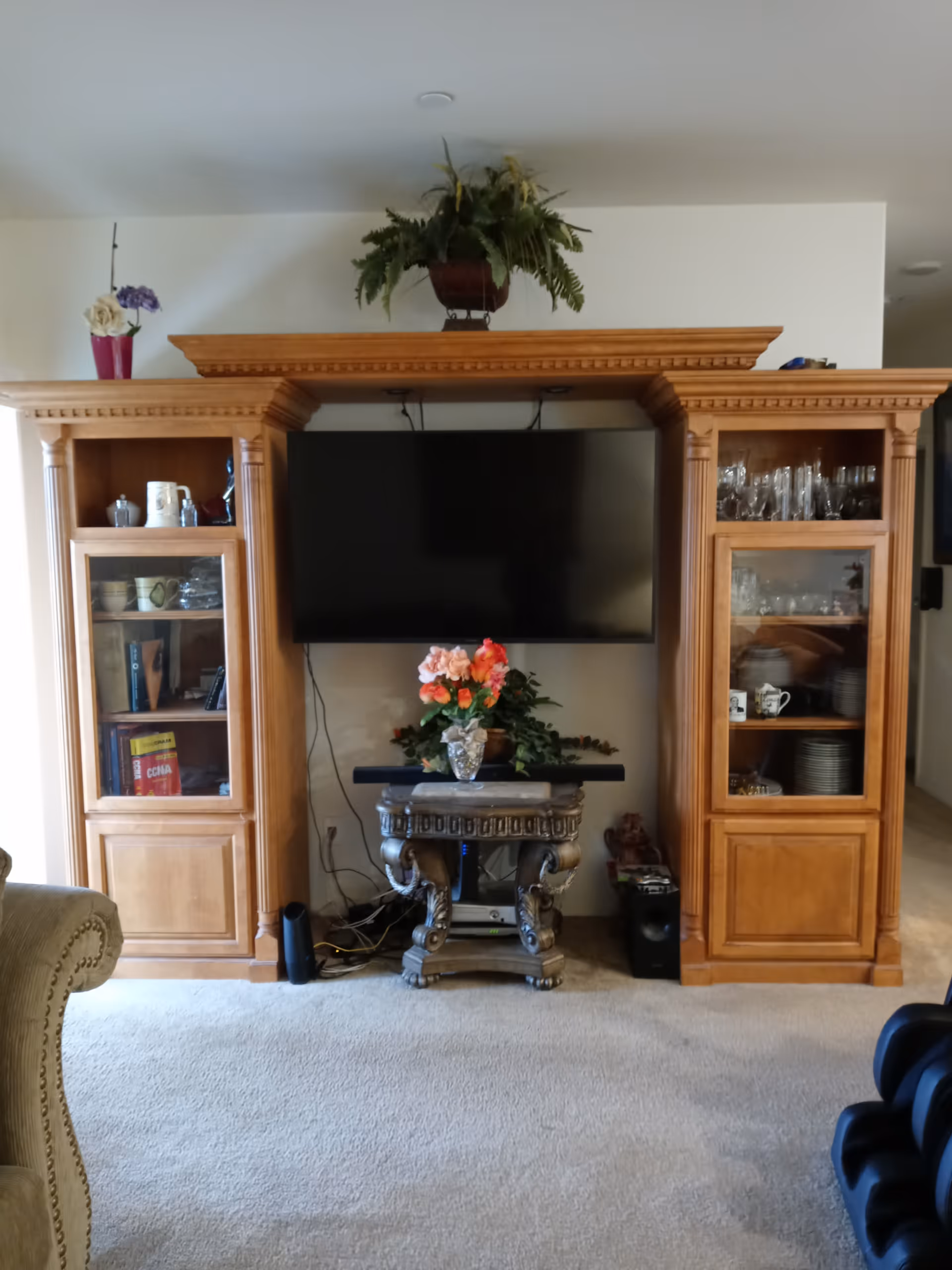A living room area featuring a large wooden entertainment center with a flat-screen TV mounted in the center. The entertainment center has glass cabinets on both sides displaying dishes, glassware, and books. Below the TV is a decorative table with a vase of colorful flowers and some electronic devices. There is a beige carpet on the floor and a potted plant on top of the entertainment center.