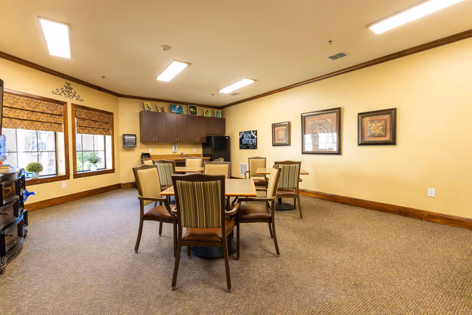 Small dining/activity room with several tables and chairs, a kitchenette with cabinets and a refrigerator, framed artwork on the walls, and windows with patterned shades.