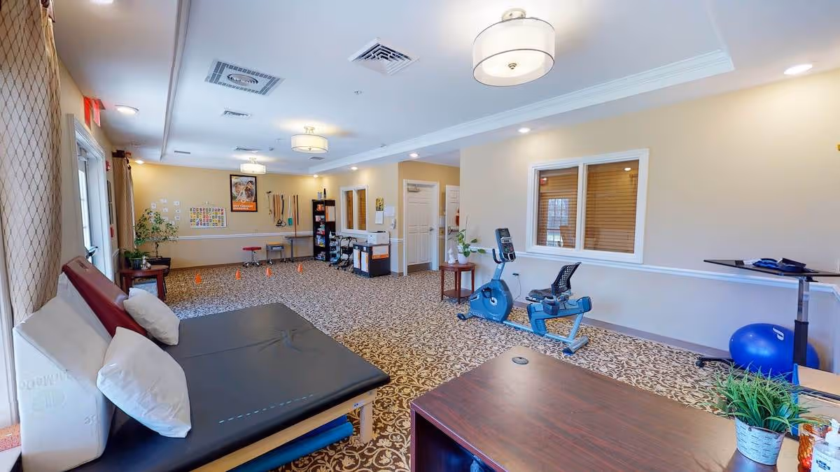 A spacious therapy or exercise room with patterned carpet, beige walls, and ceiling lights. The room contains a padded therapy table with pillows, an exercise bike, a blue exercise ball, a small table with a plant, and various therapy equipment along the far wall. There is a window with blinds on one wall and a door with a curtain on the other side.