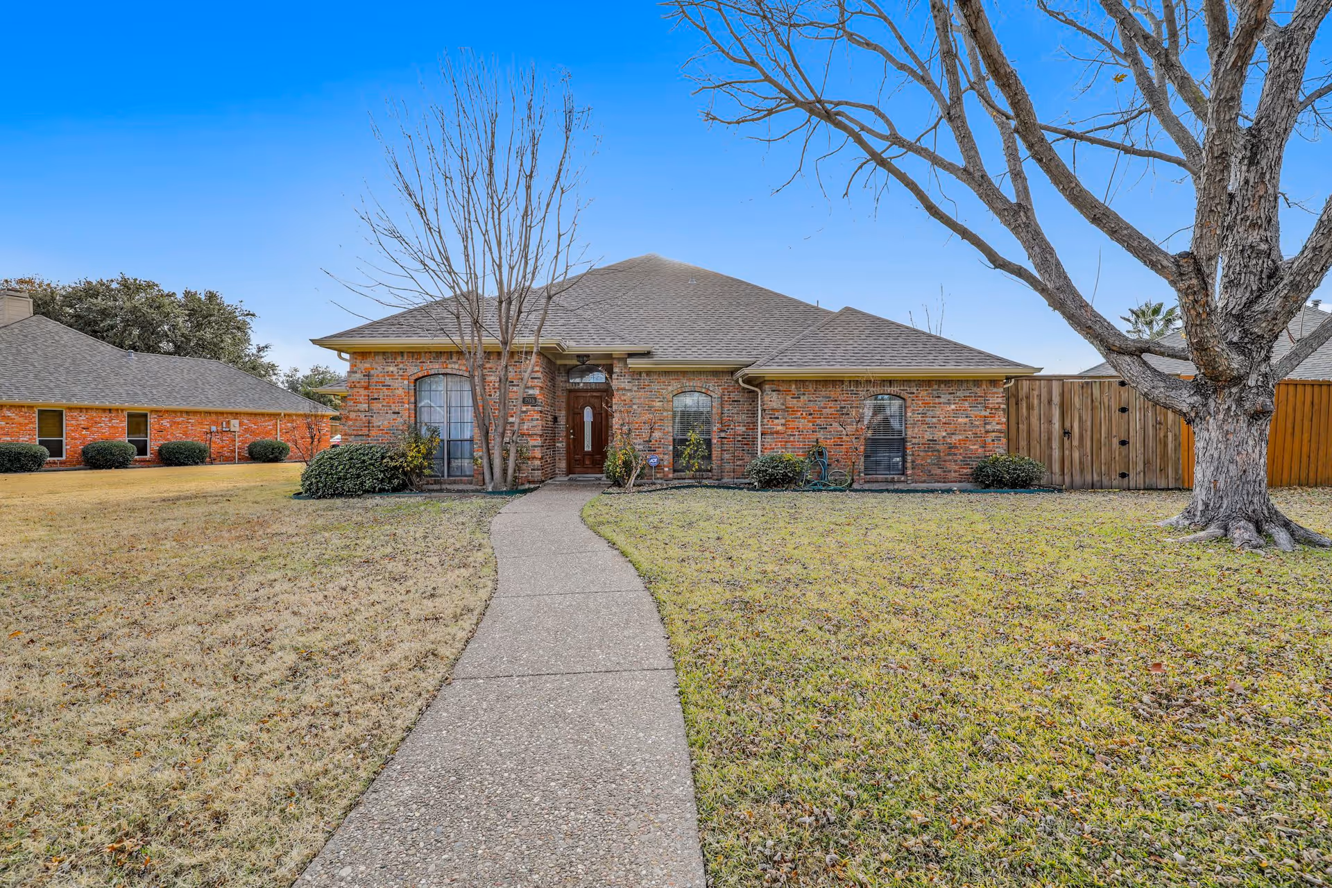 Front exterior view of a single-story brick house with a gray shingled roof, a concrete walkway leading to the front door, leafless trees, and a lawn with some bushes and grass.