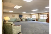A spacious common area with carpeted floor, multiple upholstered armchairs and sofas arranged around the room, a wall-mounted flat screen TV, two framed pictures on the wall, large windows with blinds, and a wooden shelving unit in the corner. The ceiling has recessed lighting panels.