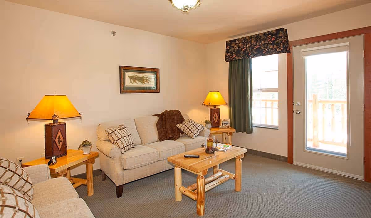 A cozy living room with two beige sofas adorned with patterned pillows and a brown throw blanket. There are two wooden side tables, each with a lamp featuring a warm yellow shade. A wooden coffee table sits in the center with a remote control, a candle, and some magazines on it. The room has a window with green curtains and a floral valance, and a glass door leading to an outdoor balcony with wooden railings. The walls are light-colored and there is a framed picture hanging above one sofa.