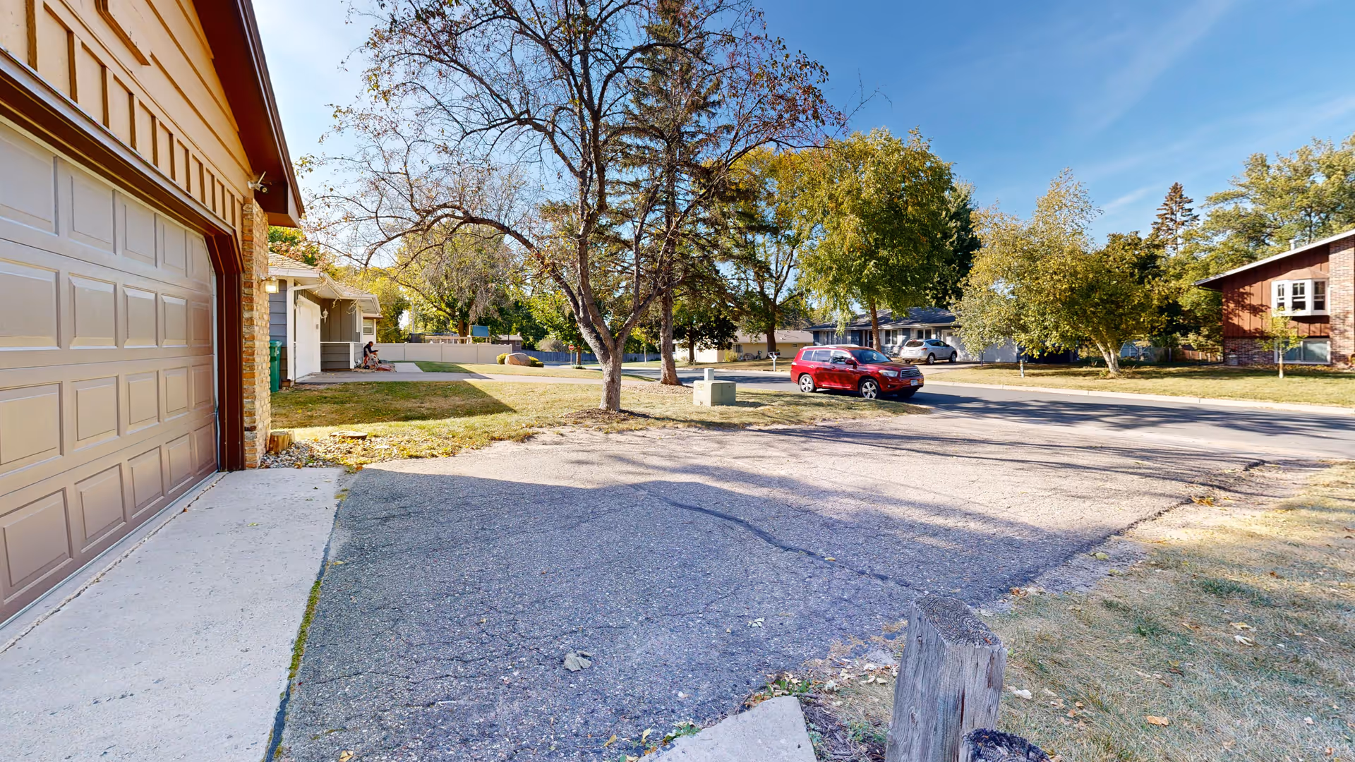 Residential driveway and garage in the foreground with trees and a red car parked on the street in a neighborhood.