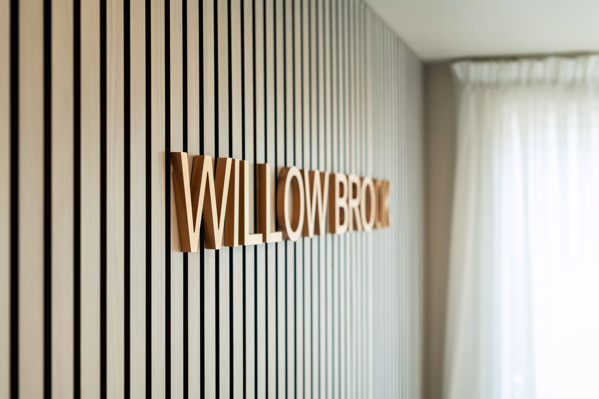 Close-up view of a wall with vertical wooden slats and large 3D letters spelling 'WILLOW BROOK' mounted on it, with a sheer white curtain and window in the background.