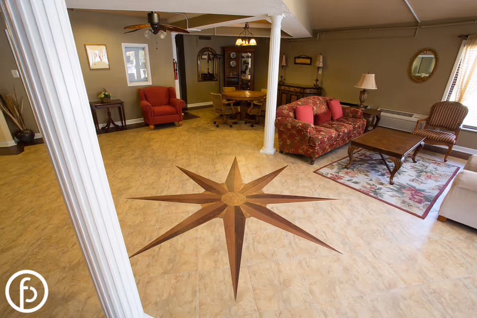 A spacious living room area with beige tiled flooring featuring a large wooden star design in the center. The room includes a floral patterned sofa with red cushions, a wooden coffee table on a floral rug, a striped armchair near a window with blinds, a red armchair, a round wooden table with chairs, and various decorative items including lamps, mirrors, and framed pictures on beige walls.