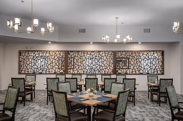 A dining room with multiple square wooden tables set with blue placemats, glasses, and small flower arrangements. The chairs have green patterned upholstery. The room features decorative wooden lattice panels on the far wall and modern chandeliers hanging from the ceiling. The carpet is patterned in shades of gray.