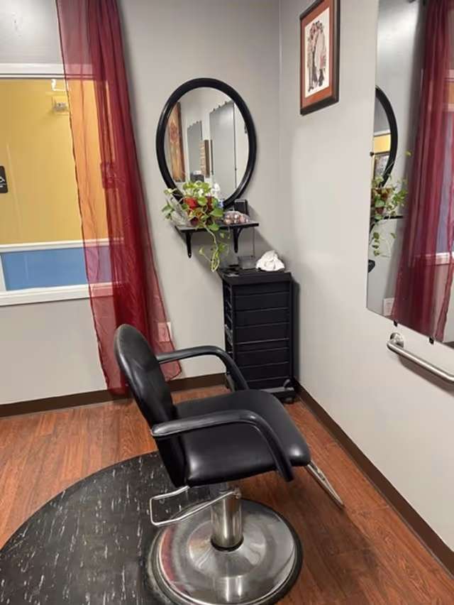 Black salon chair in front of a round wall mirror, red curtains, and a small shelf with a potted plant in an interior room.
