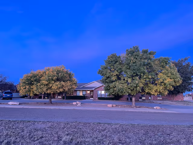 Single-story brick building with large trees and a lawn in front, seen from across the street under a vivid blue sky.