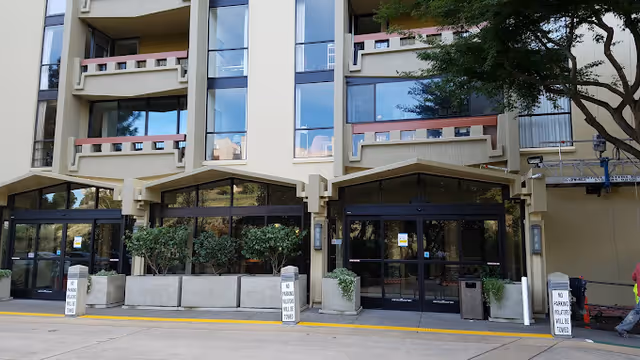 Front entrance of a multi-story senior living building with glass doors, planters, and balconies above.
