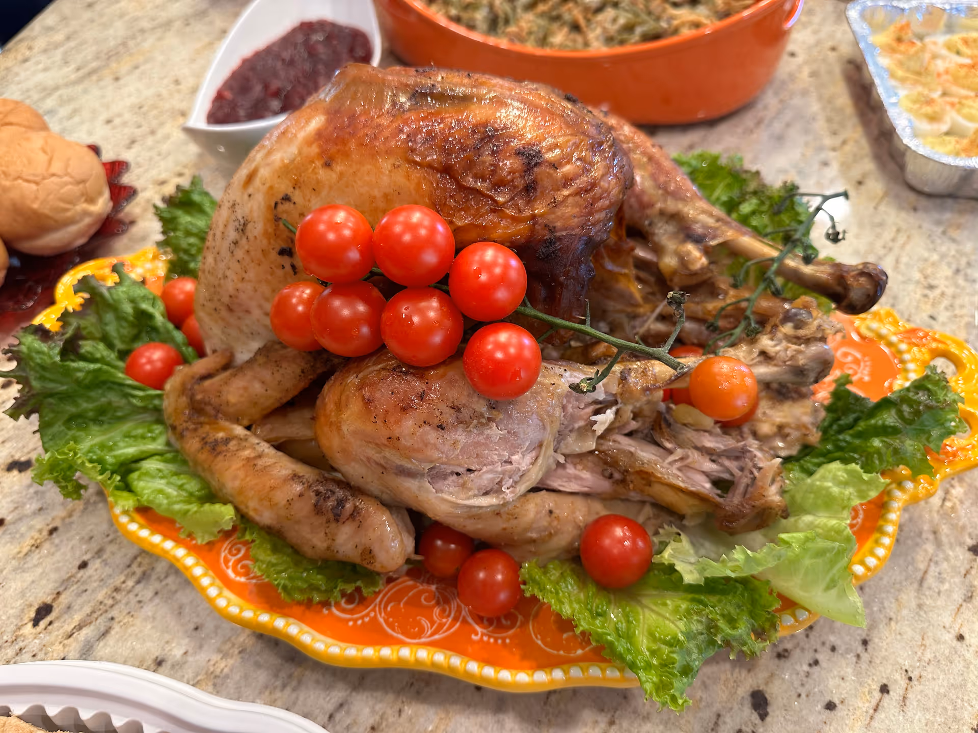 A roasted whole turkey garnished with cherry tomatoes and leafy greens on an orange decorative platter, surrounded by side dishes including cranberry sauce, rolls, and other food items on a countertop.
