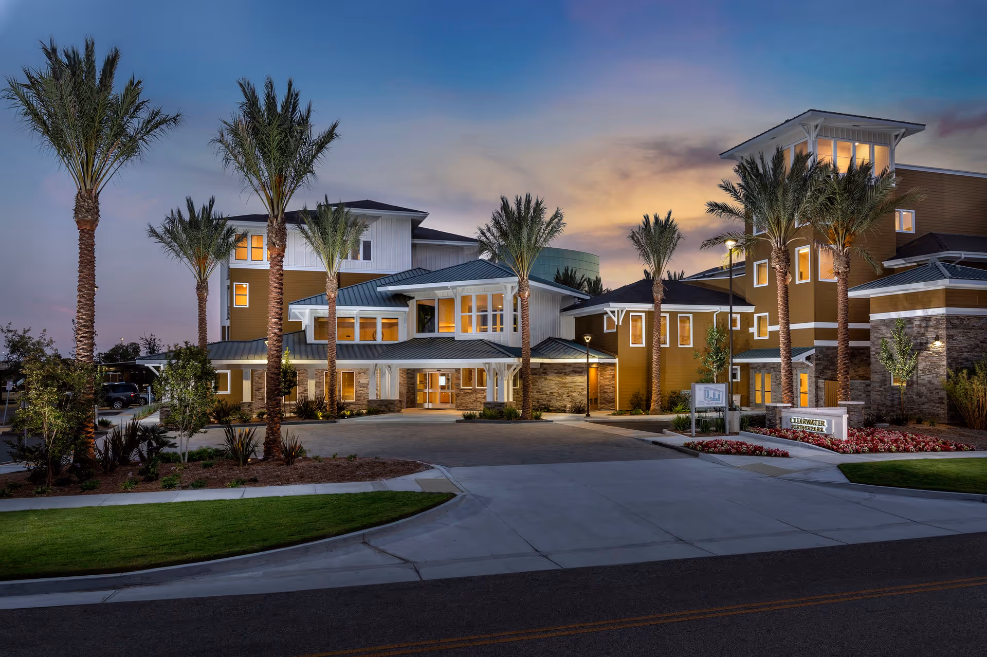 Exterior view of Clearwater at Riverpark facility at dusk, showing a modern multi-story building with large windows, palm trees, landscaped greenery, and a driveway entrance.