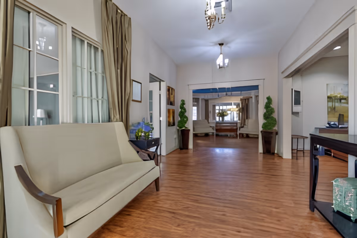 A spacious interior hallway with wooden flooring, beige walls, and ceiling lights. On the left side, there is a cream-colored sofa with wooden armrests next to large windows with beige curtains. The hallway leads to another room with more seating and decorative plants in tall pots on either side of the entrance.